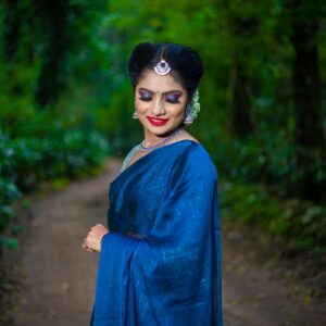Woman in traditional blue saree standing gracefully amidst a vibrant green forest.