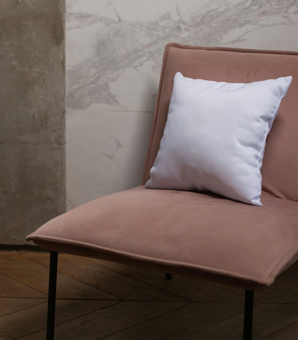 Beige chair with a white pillow in a minimalist room with wooden flooring and marble wall.